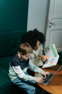 A woman tutors a child with a laptop and book, fostering learning in a cozy setting.